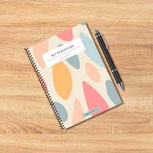 May include: A spiral-bound journal with a cream-colored cover featuring abstract shapes in pink, orange, and blue. The journal has the text "MY SCRIPTURE journal" and a black pen rests beside it. The journal is from Ember Design & Co.