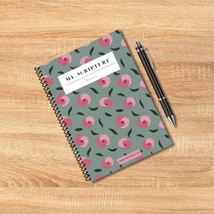 May include: Spiral-bound notebook with a floral pattern of pink roses and green leaves on a teal background. The notebook has the text "MY SCRIPTURE Notes" and a black pen is next to it. The notebook is on a wooden surface.