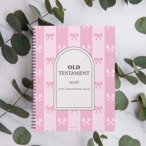 May include: A spiral-bound study journal with a pink bow pattern. The cover features the text "OLD TESTAMENT 2026 Come, Follow Me Study Journal" within a white arched frame. Green foliage accents the journal.