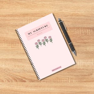 May include: A spiral-bound notebook with a light pink cover featuring the text "MY SCRIPTURE Notes" in a rectangular box, and a row of pink flowers. A black pen rests on the notebook. The notebook is on a wooden surface.