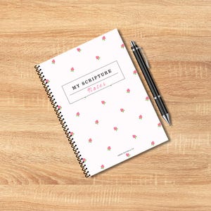 May include: A spiral-bound notebook with a white cover featuring a pattern of small pink roses. The text "MY SCRIPTURE Notes" is displayed in a rectangular box. A black pen rests on the notebook. The notebook is on a wooden surface.