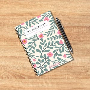 May include: A spiral-bound journal with a floral design featuring pink flowers and green leaves on an off-white background. The journal has the text "MY SCRIPTURE Journal" and a black pen rests beside it.