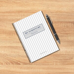 May include: A spiral-bound notebook with a gray and white striped cover and the text "MY SCRIPTURE Notes" in a gray rectangle. A black pen rests on the right side of the notebook. The notebook is on a wooden surface.