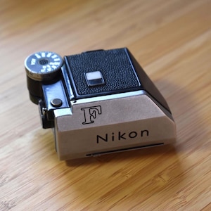 May include: A black and silver Nikon F camera prism viewfinder attachment. The device features a textured black top section, a silver metal front plate with the engraved letter F and the Nikon brand name, and a circular dial on the side.
