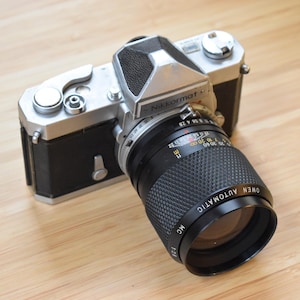 Pode incluir: Uma câmera Nikkormat vintage com corpo preto e prateado e lente preta. A lente tem a inscrição "OWEN AUTOMATIC". A câmera está posicionada sobre uma superfície de madeira marrom clara.