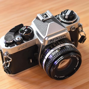 May include: A silver and black Nikon FE2 35mm film camera with a 50mm lens. The camera has a textured top plate and a black leather grip. The lens has a focus ring and aperture settings. The camera is on a wooden surface.