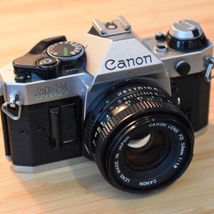 Könnte beinhalten: Canon AE-1 Filmkamera mit schwarzem Gehäuse und silberner Oberseite. Das Objektiv ist ein Canon Lens FD 50mm 1:1.8. Auf der Kamera sind die Texte "Canon" und "AE-1" zu sehen. Die Kamera liegt auf Holz.