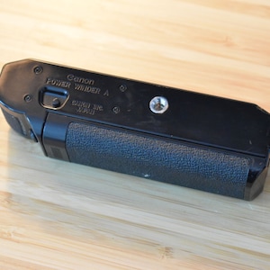 Puede incluir: Accesorio de cámara Canon Power Winder A negro con agarre texturizado. El texto "Canon Power Winder A" y "Canon Inc. Japan" son visibles. El accesorio es rectangular con bordes redondeados.