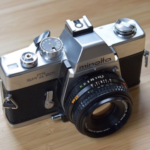 May include: A silver and black Minolta SRT 201 35mm film camera featuring a black MD Rokkor-X 45mm 1:2 lens. The camera body includes a top-mounted shutter speed dial, a film advance lever, a hot shoe, and a textured black grip on the sides.
