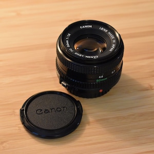 Op de afbeelding: Een Canon 50mm f/1.8 lens met een zwarte lensdop. De lens is zwart met groene en witte markeringen, waaronder "50mm" en "1:1.8". De lensdop heeft het Canon-logo. De lens ligt op een houten oppervlak.