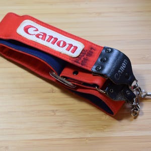 Puede incluir: Correa de cámara Canon roja y azul con el logotipo de Canon en blanco. La correa tiene extremos de cuero negro y herrajes metálicos para la fijación. La correa está sobre una superficie de madera.