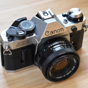 Könnte beinhalten: Eine silberne und schwarze Canon AE-1 Program 35mm Filmkamera mit einem schwarzen Objektiv. Die Kamera hat das Canon-Logo und den Text "AE-1 PROGRAM" auf der Vorderseite. Das Objektiv ist mit "50mm 1:1.8" und "MADE IN JAPAN" gekennzeichnet.
