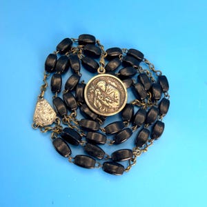 May include: A rosary featuring smooth, rectangular black beads linked by a brass-toned metal chain. A circular metal pendant depicts Saint Anthony holding a child, with the text SAINT ANTHONY inscribed along the upper edge of the medal.