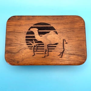 May include: A rectangular wooden box with rounded corners. The box features a laser-etched design of a camel silhouette within a circular frame, set against a striped background. The wood has a warm, brown tone.