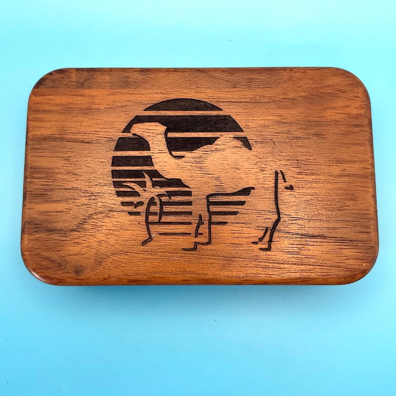 May include: A rectangular wooden box with rounded corners. The box features a laser-etched design of a camel silhouette within a circular frame, set against a striped background. The wood has a warm, brown tone.