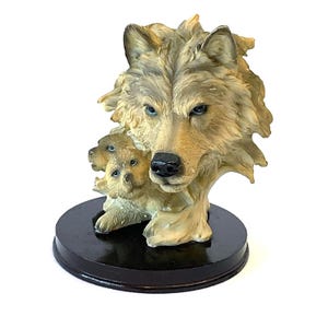 May include: A ceramic figurine of a wolf with two pups. The wolf is light brown and gray with black eyes. The pups are light brown with black eyes. The figurine is on a black wooden base.