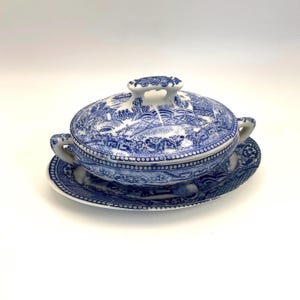 May include: A blue and white ceramic soup tureen with a matching saucer. The tureen has a lid with a handle and two side handles. The design features a detailed landscape scene. The set is made of ceramic.
