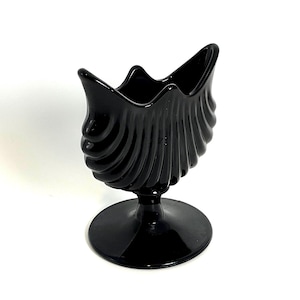 May include: A black, vintage-style glass vase with a shell-like design. The vase has a pedestal base and a flared, scalloped top. The exterior features vertical, curved ridges. The vase is made of glossy black glass.