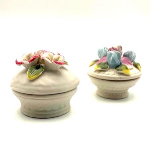 Könnte beinhalten: Zwei kleine, dekorative Schmuckdosen aus Keramik. Eine zeigt rosa und weiße Blumen mit grünen und gelben Akzenten, die andere blaue, rosa und gelbe Blumen. Beide Dosen sind cremefarben mit einem floralen Design.