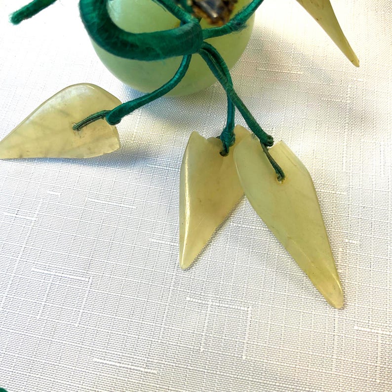 K&ouml;nnte beinhalten: Nahaufnahme eines dekorativen Jade-Ornaments. Das Ornament hat eine hellgr&uuml;ne, runde Basis mit gr&uuml;nem Faden und mehrere hellgelbe, blattf&ouml;rmige Teile. Die Bl&auml;tter sind mit gr&uuml;nem Faden an der Basis befestigt. Der Hintergrund ist eine wei&szlig;e, strukturierte Oberfl&auml;che.