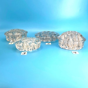 May include: Four clear glass flower frogs of varying shapes and sizes, each featuring multiple circular openings for stems. The items are labeled with small white paper tags numbered one through four, arranged on a solid blue surface.