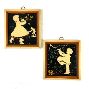 May include: Two framed silhouette art pieces. One shows a child holding flowers with another figure. The other depicts a child playing with a ball under a tree. Both are in light wood frames with a dark background.