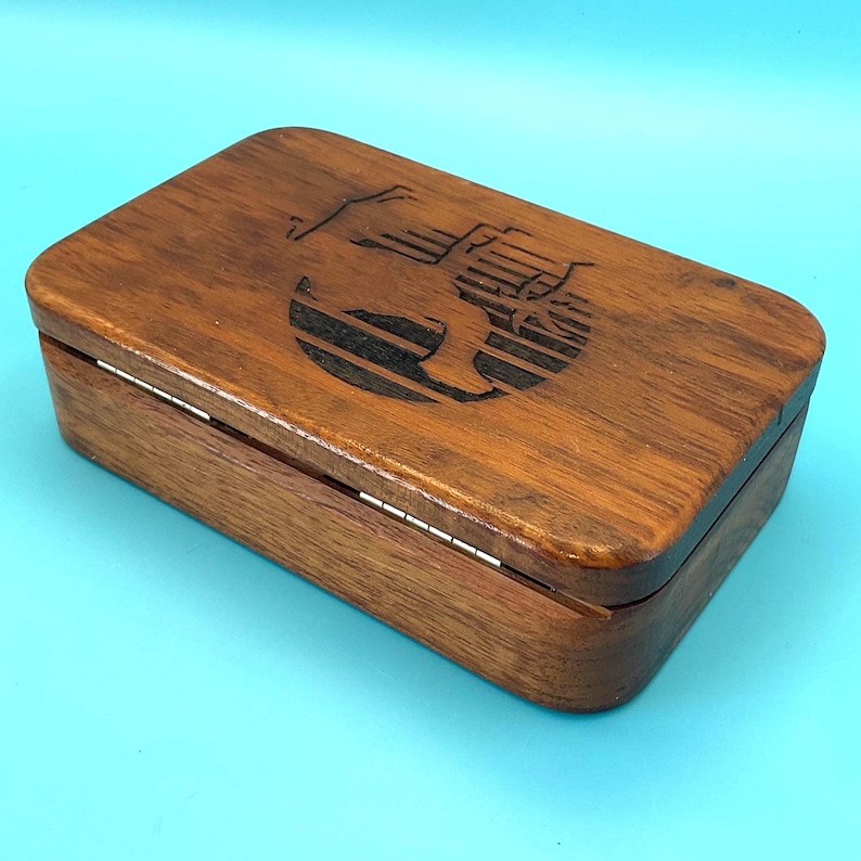 May include: A rectangular wooden box with a hinged lid. The box is made of dark brown wood and features a black engraved design on the top. The box is on a light blue surface.