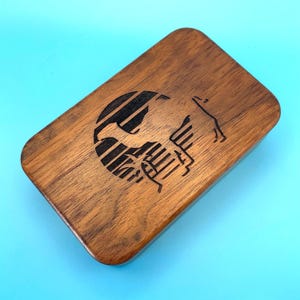 May include: A rectangular wooden box with rounded edges. The box is made of a dark brown wood and features a black graphic design on the top. The design appears to be a stylized image of a horse and a globe.