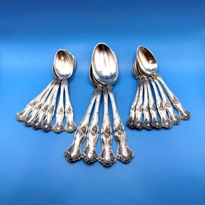 May include: A collection of silver-toned metal spoons arranged in three distinct groups. Each spoon features an ornate handle with a raised grape and vine motif. The set includes various sizes of spoons, all displaying a polished metallic finish.
