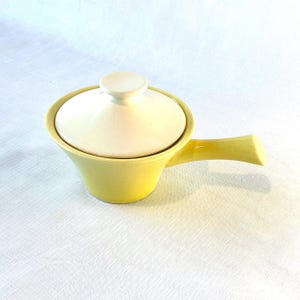 Op de afbeelding: Een gele keramische serveerschaal met een wit deksel en een handvat. De schaal is perfect om sauzen, dips of andere kleine gerechten te serveren.