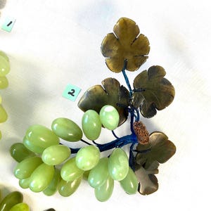 K&ouml;nnte beinhalten: Eine dekorative Weinreben-Skulptur mit gr&uuml;nen Trauben und braunen Bl&auml;ttern. Die Trauben sind oval geformt und unterschiedlich gro&szlig;. Die Bl&auml;tter sind dunkelbraun. Der Stiel ist dunkelblau. Die Skulptur befindet sich auf wei&szlig;em Hintergrund.