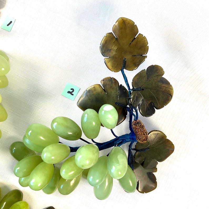 K&ouml;nnte beinhalten: Eine dekorative Weinreben-Skulptur mit gr&uuml;nen Trauben und braunen Bl&auml;ttern. Die Trauben sind oval geformt und unterschiedlich gro&szlig;. Die Bl&auml;tter sind dunkelbraun. Der Stiel ist dunkelblau. Die Skulptur befindet sich auf wei&szlig;em Hintergrund.