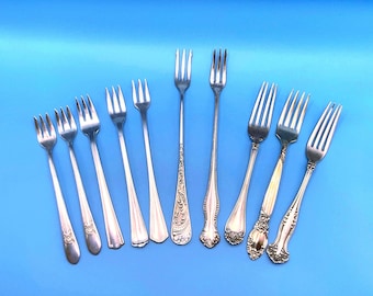 May include: A collection of antique silver forks with various handle designs. The forks are arranged on a blue background, showcasing their tines and decorative patterns. The collection includes forks with ornate and simple designs.