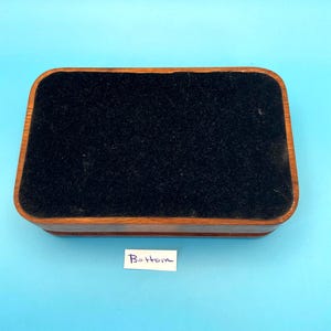 May include: A rectangular wooden box with a dark brown exterior and a black velvet interior. The box has rounded corners and a label that reads "Bottom". The box is set against a light blue background.