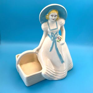 May include: A vintage ceramic figurine of a woman in a white dress and a blue hat, holding a small bouquet. The figurine has a basket attached to the side. The dress has blue ribbon accents. The figurine is on a blue background.