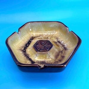 May include: A hexagonal ceramic ashtray featuring a glossy glaze in shades of honey yellow and dark brown. The center of the basin displays a raised, embossed geometric pattern consisting of six triangular segments arranged in a star shape.