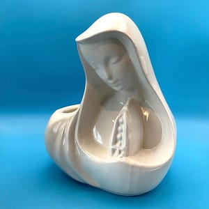 May include: A white ceramic figure of a woman in a draped head covering, with her head bowed. The figure has a serene expression and is holding her hands in prayer. The figurine has a smooth, glossy finish and is set against a blue background.