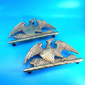 May include: A pair of polished brass eagle door knockers or decorative hardware pieces. Each features a spread-winged eagle clutching a shield with three stars and vertical stripes, perched atop a horizontal bar with leaf and arrow motifs.