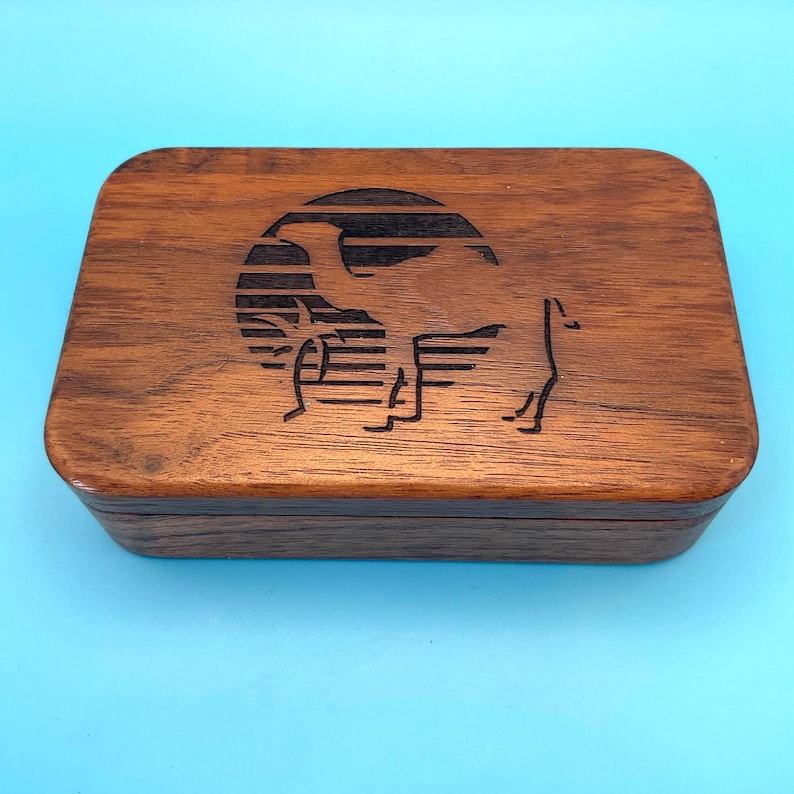 May include: A rectangular wooden box with a camel silhouette design etched on the lid. The camel is set against a circular background with horizontal lines, all in a dark brown color. The box has rounded edges and a smooth finish.