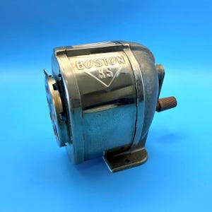 以下が含まれることがあります： 光沢のあるクロームメッキの金属製ボディとダークグレーのベースを備えた、ボストンKSのヴィンテージ手動鉛筆削り。前面には三角形の輪郭の中に「BOSTON KS」という浮き彫りの文字が刻まれています。側面には小さな茶色のプラスチック製クランクハンドルが付いています。