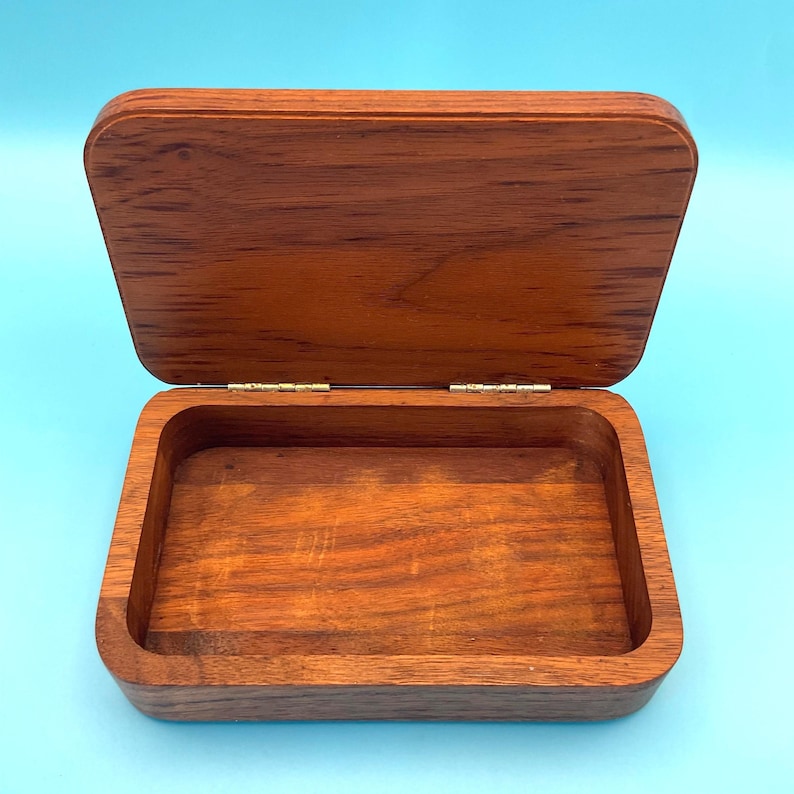 May include: Open rectangular wooden box with a hinged lid. The box is made of a reddish-brown wood, with visible grain patterns. The lid is slightly ajar, revealing the empty interior. The box is set against a light blue background.