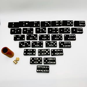 Pode incluir: Um conjunto de dominós pretos com pontos brancos, dispostos sobre uma superfície branca. Um suporte de dominó de madeira e dois dados também estão presentes. Os dominós são retangulares e apresentam diferentes números de pontos.