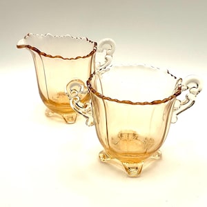 May include: A vintage amber glass creamer and sugar bowl set. The creamer has a spout and a scalloped rim. The sugar bowl has two handles and a matching scalloped rim. Both pieces sit on three small feet.