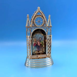 May include: A gold-toned metal triptych featuring an arched gothic frame with open side panels. The central image depicts the Holy Family, including a figure in a blue head covering, a central child, and a figure in green robes, all set on a base.