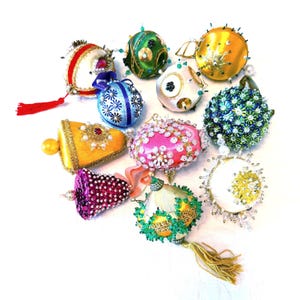 Puede incluir: Una colección de doce adornos decorativos de diversas formas, incluyendo esferas, campanas y óvalos. Estos artículos presentan telas de satén vibrantes, abalorios intrincados, lentejuelas, adornos de metal dorado y borlas de colores.