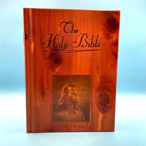 May include: A wooden-covered Holy Bible with the title in elegant script. The cover features a portrait of a man with long hair and a beard. The wood has a reddish-brown hue.