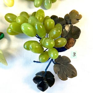 K&ouml;nnte beinhalten: Ein dekorativer Haufen gr&uuml;ner Trauben mit dunkelgr&uuml;nen Bl&auml;ttern und blauen Stielen. Die Trauben sind oval geformt und scheinen aus einem glatten, polierten Material zu bestehen. Die Bl&auml;tter haben detaillierte Adern. Ein kleines wei&szlig;es Etikett mit der Nummer 3 ist sichtbar.