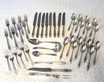 May include: A large collection of silver-toned stainless steel flatware including forks, knives, and spoons. The set features ornate decorative detailing on the handles of the knives, spoons, and serving pieces, with plain tines on the forks.
