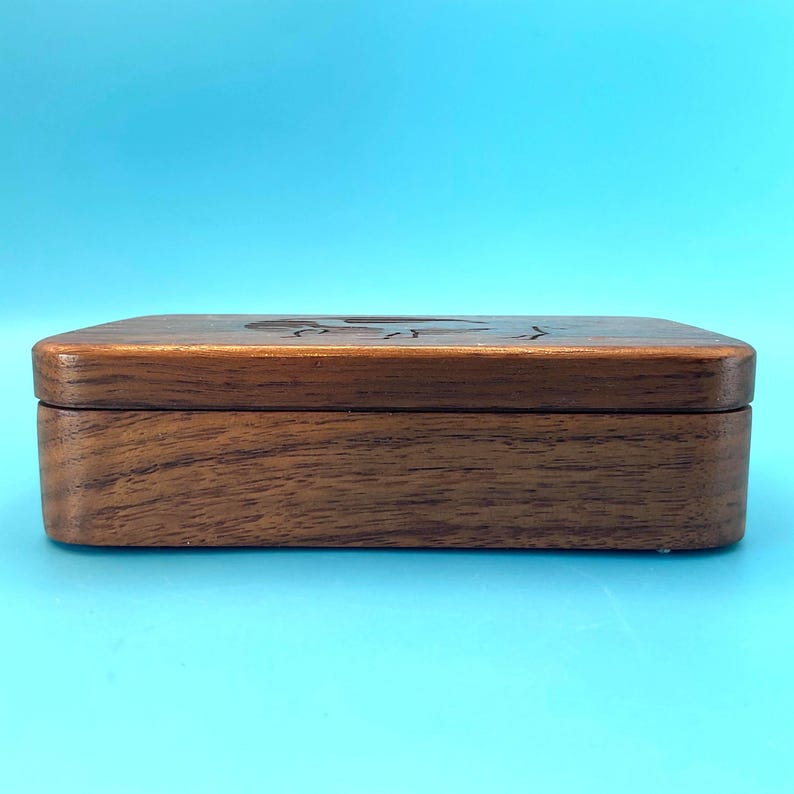 May include: A rectangular wooden box with a dark brown finish. The box has a lid with a carved design on top. The box is sitting on a light blue surface.