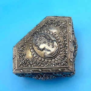 May include: Silver-toned, antique jewelry box with a cameo design. The box features a raised relief of a woman's profile within an oval frame, surrounded by floral and leaf motifs. The box has a rectangular shape with beveled edges.